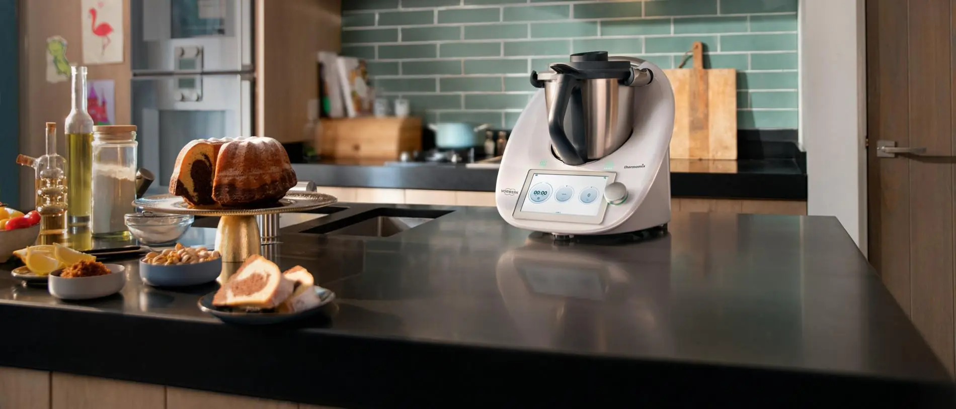 Hochwertige Küchenarbeit mit Thermomix-ähnlichem Multikocher auf Kücheninsel, daneben Kuchenstücke und Snacks auf Tellern.