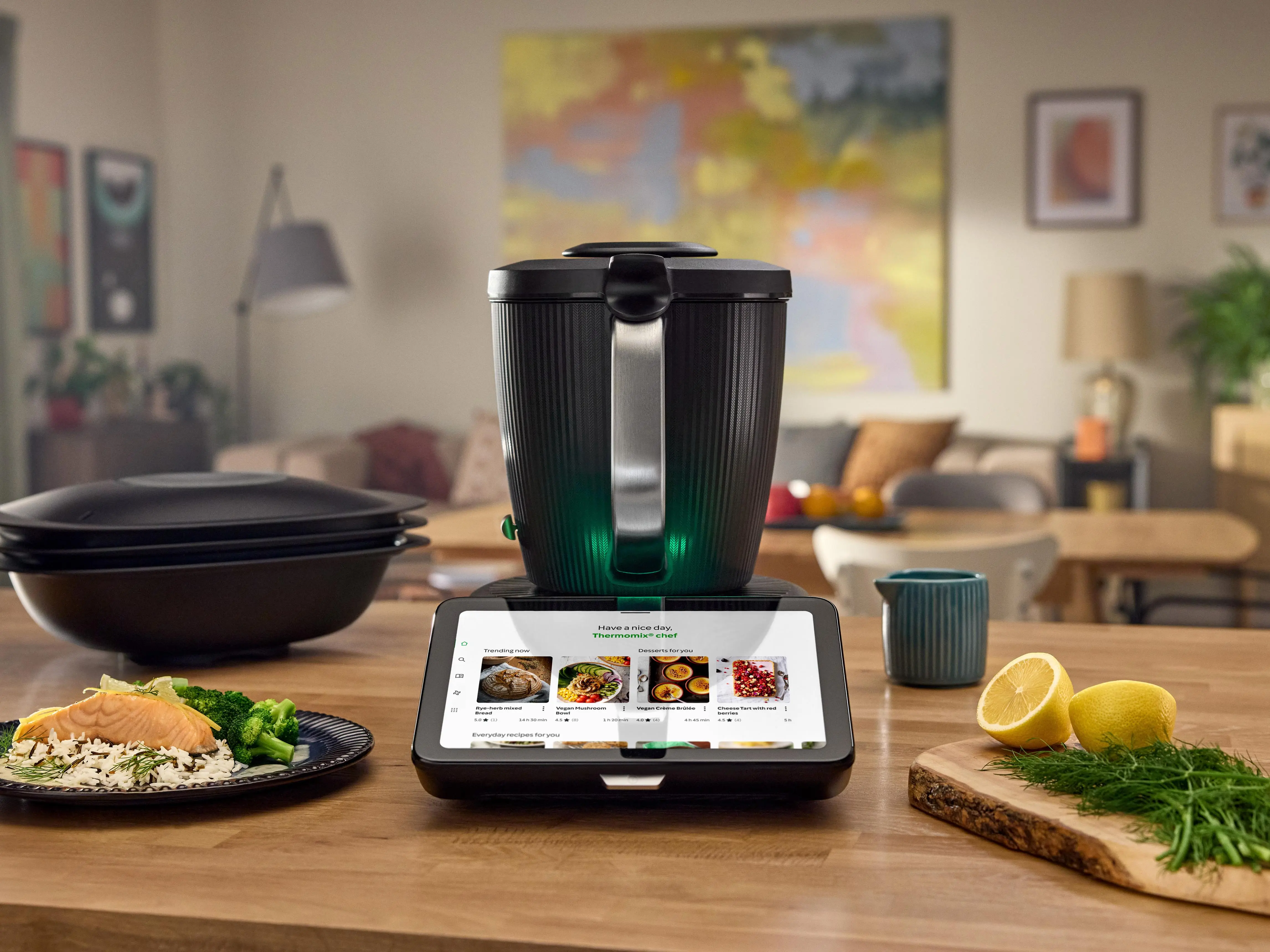 Multifunktionale Küchenmaschine am Küchentisch mit Tablet-Bedienoberfläche, daneben Teller mit Essen, Zitronen und Kräutern.