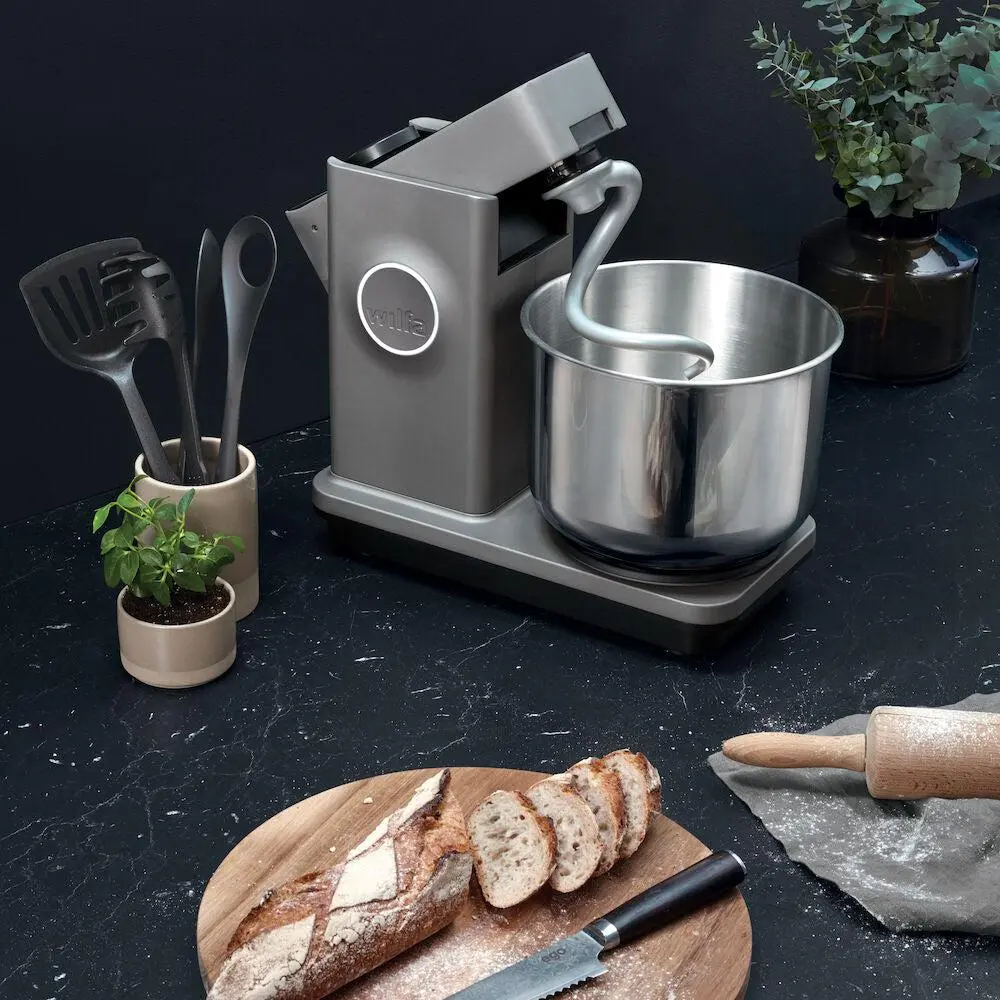 Küchenmaschine mit Edelstahltopf auf Küchenarbeitsplatte, Brotlaib und Rollholz daneben; Küchenutensilien im Hintergrund.
