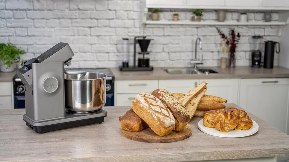 Küchenmaschine von Nella mit Edelstahl-Schüssel neben Brote und Croissants auf Holzplatte auf hellem Küchenblock.