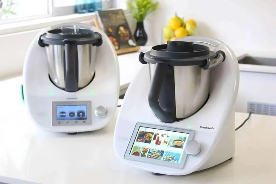 Zwei weiße Thermomix-ähnliche Küchenmaschinen mit Touchscreen-Displays auf glatter Küchenarbeitsplatte, daneben Obst im Hintergrund.